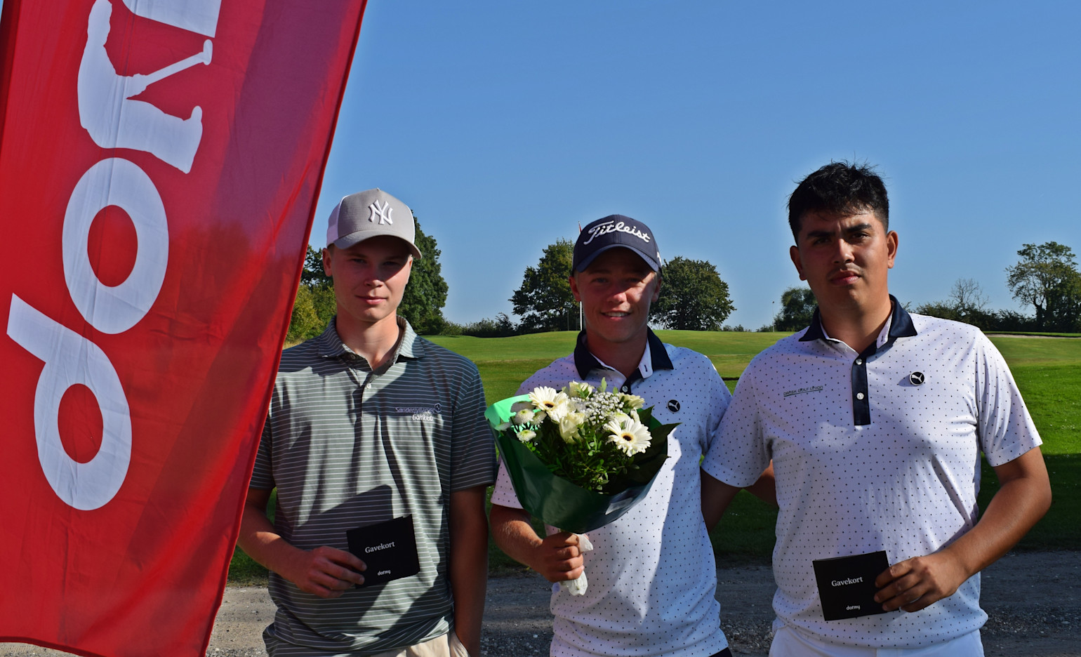 Tre unga män poserar utomhus på en golfbana, två håller presentkort och en håller en blombukett, bredvid en röd flagga.