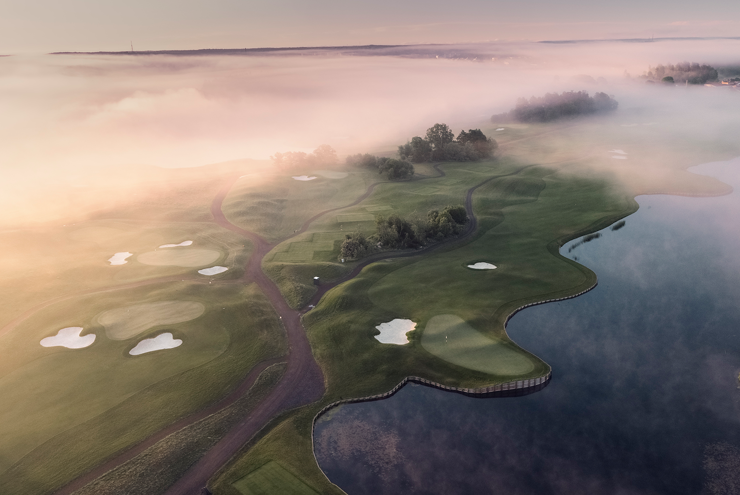 Golfbana med gröna fairways och sandbunkrar slingrar sig längs en sjö, omgiven av dimma i gryningen.
