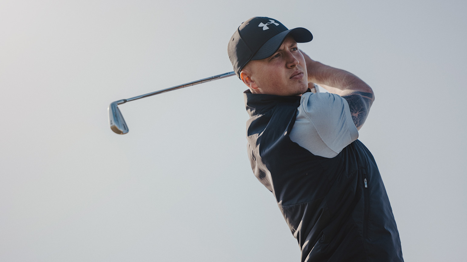 A man in a cap and golf attire swings a golf club outdoors against a bright sky.