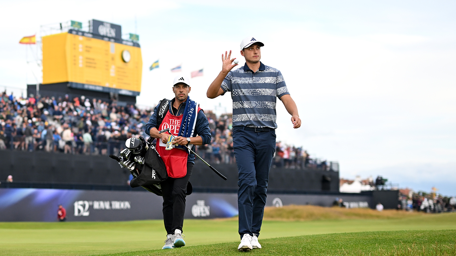 Golfspelare vinkar och håller en golfboll, caddie bär golfbag bredvid, publik och resultattavla i bakgrunden på golfbana.