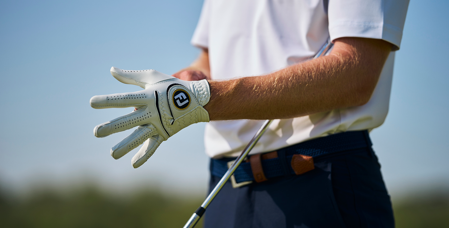 Golfspelare tar på sig en vit golfhandske med logotyp, håller en golfklubba, utomhus på golfbana.