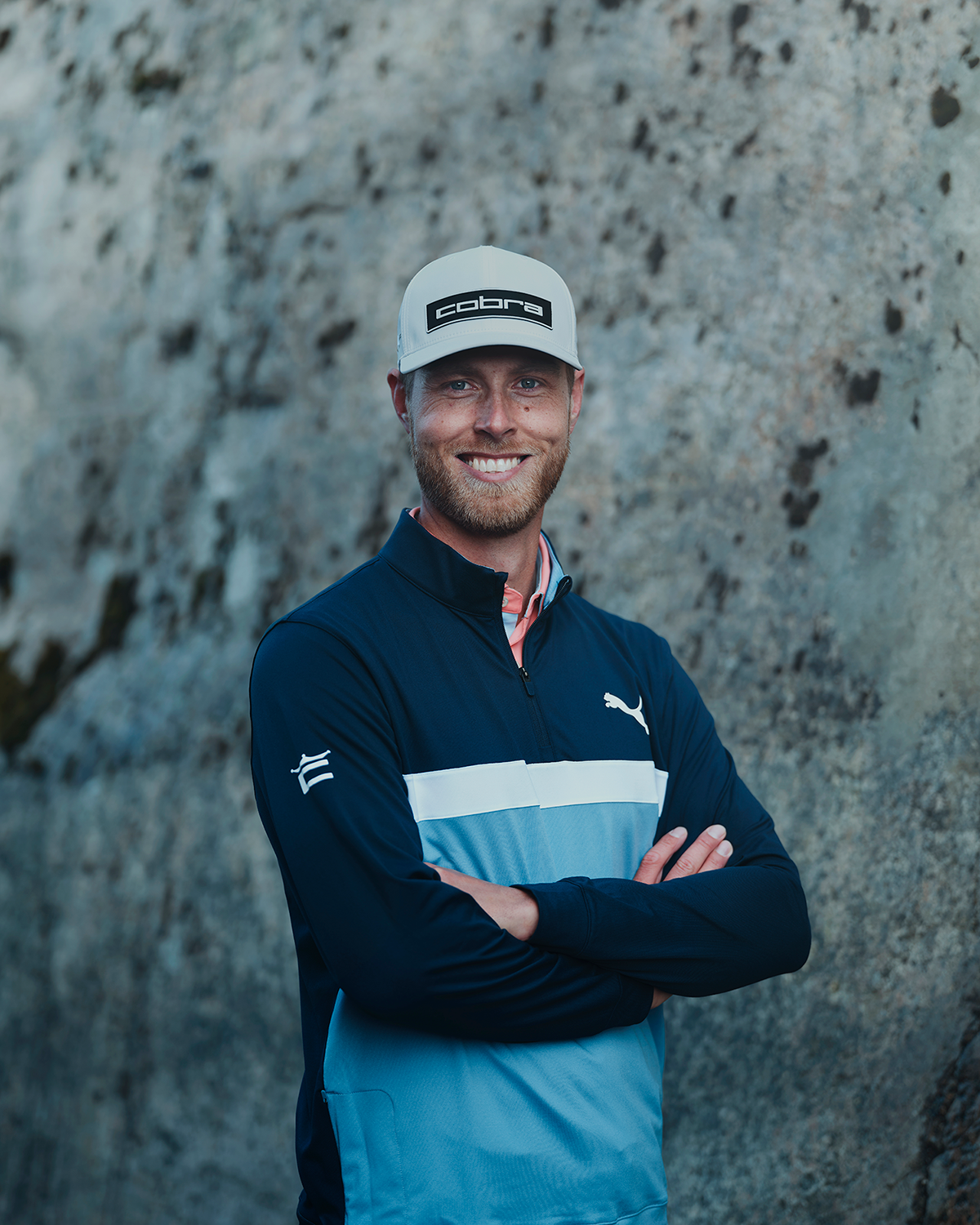 Leende man med keps med texten "cobra" och blå sportjacka med Puma-logotyp står med armarna i kors framför en stenmur.