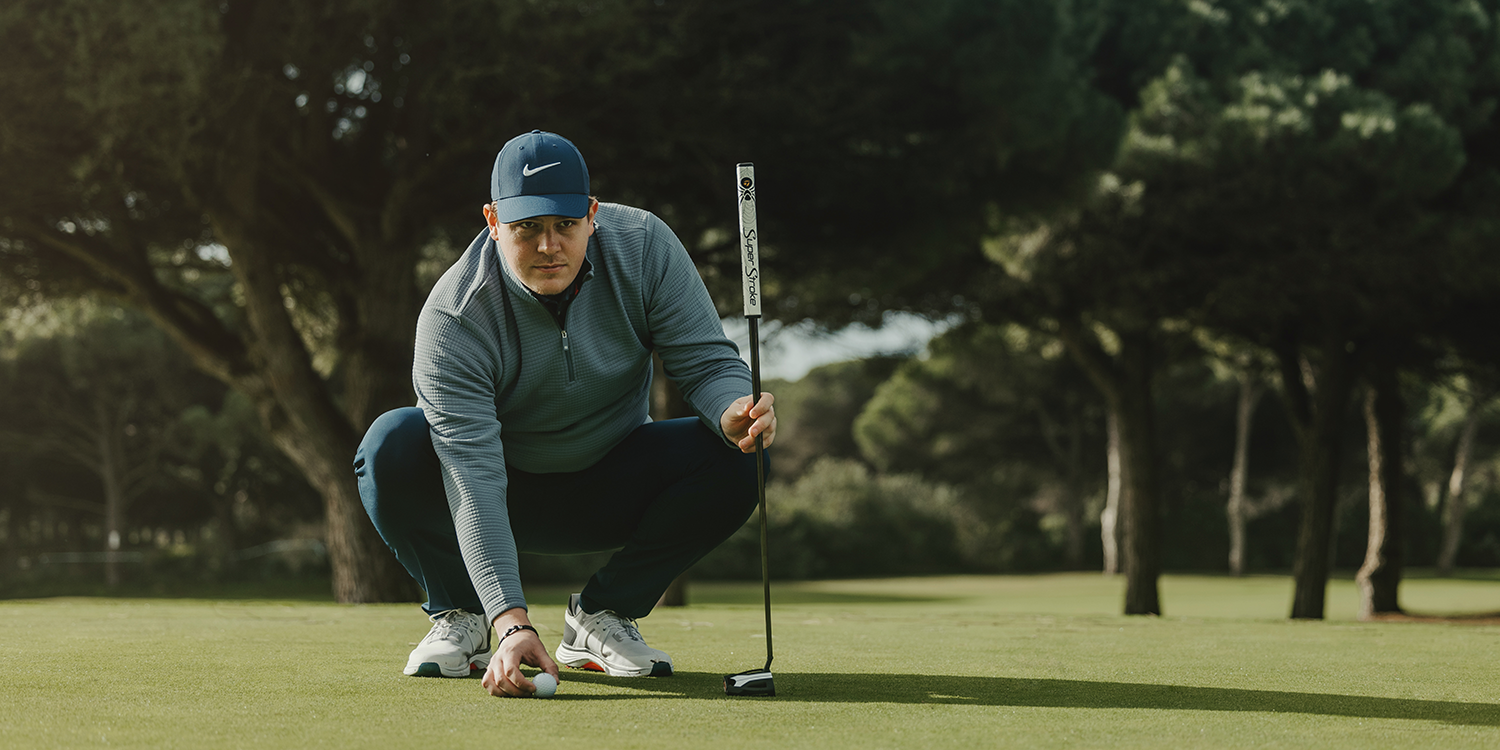 A man in a blue cap and golf attire is crouching down and placing a golf ball on the green with a putter in hand, surrounded by trees.