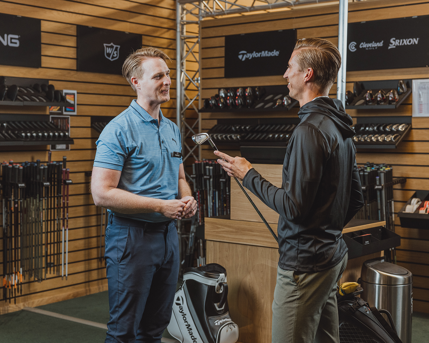 Två män samtalar i en golfbutik, där en av dem håller i en golfklubba och omges av golfutrustning och klubbor.