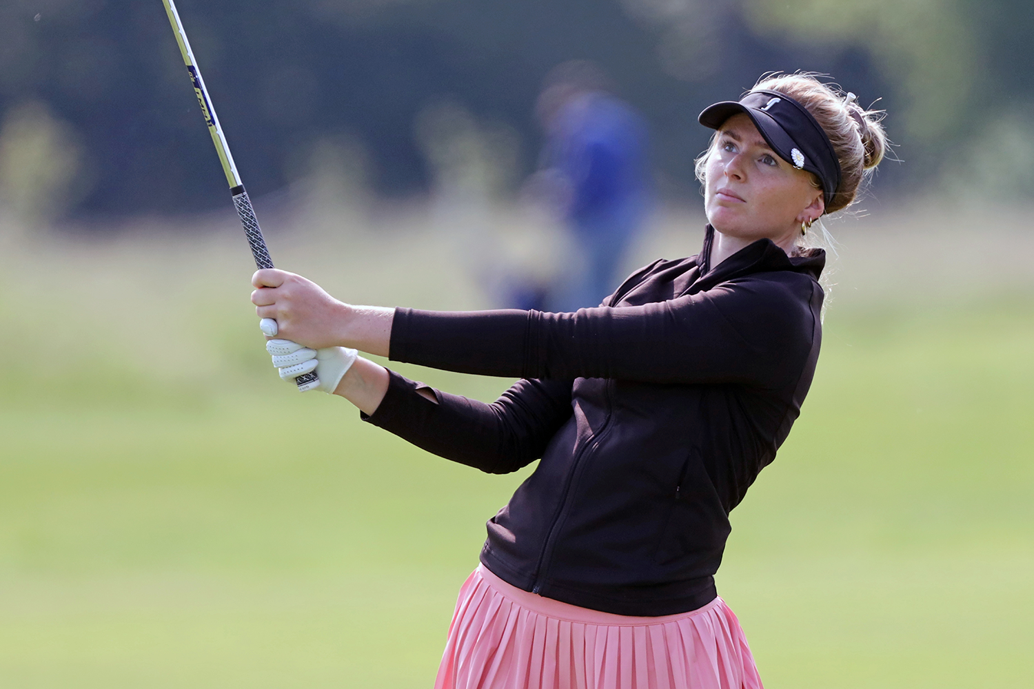 Kvinna i svart jacka och rosa kjol svingar en golfklubba på en golfbana under en solig dag.