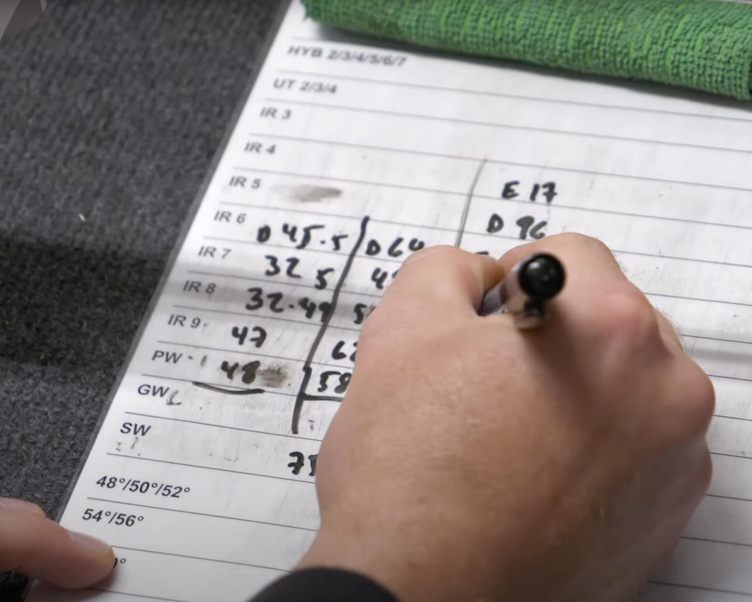 Hand skriver siffror och bokstäver med penna på ett papper med golfklubbspecifikationer och tabeller på ett skrivbord.