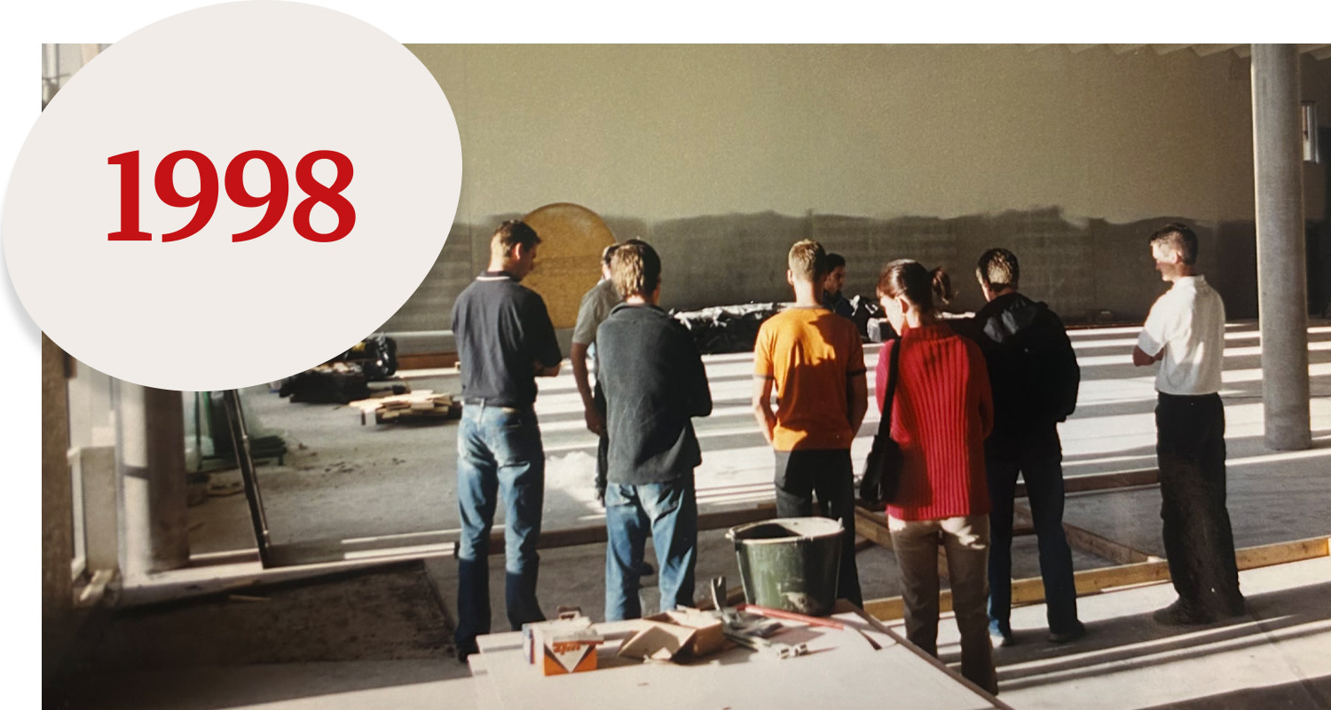 Seven people are standing in a half-finished building, looking towards a wall, with the text "1998" in red in the top left corner.