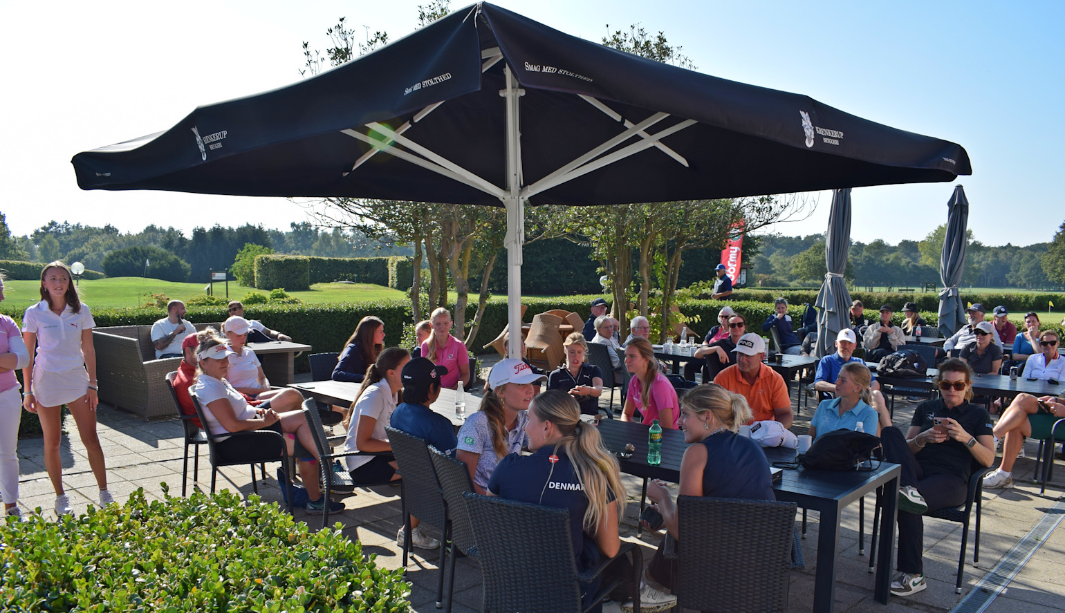 En grupp människor sitter och pratar vid bord under ett stort parasoll på en uteservering vid en golfbana en solig dag.