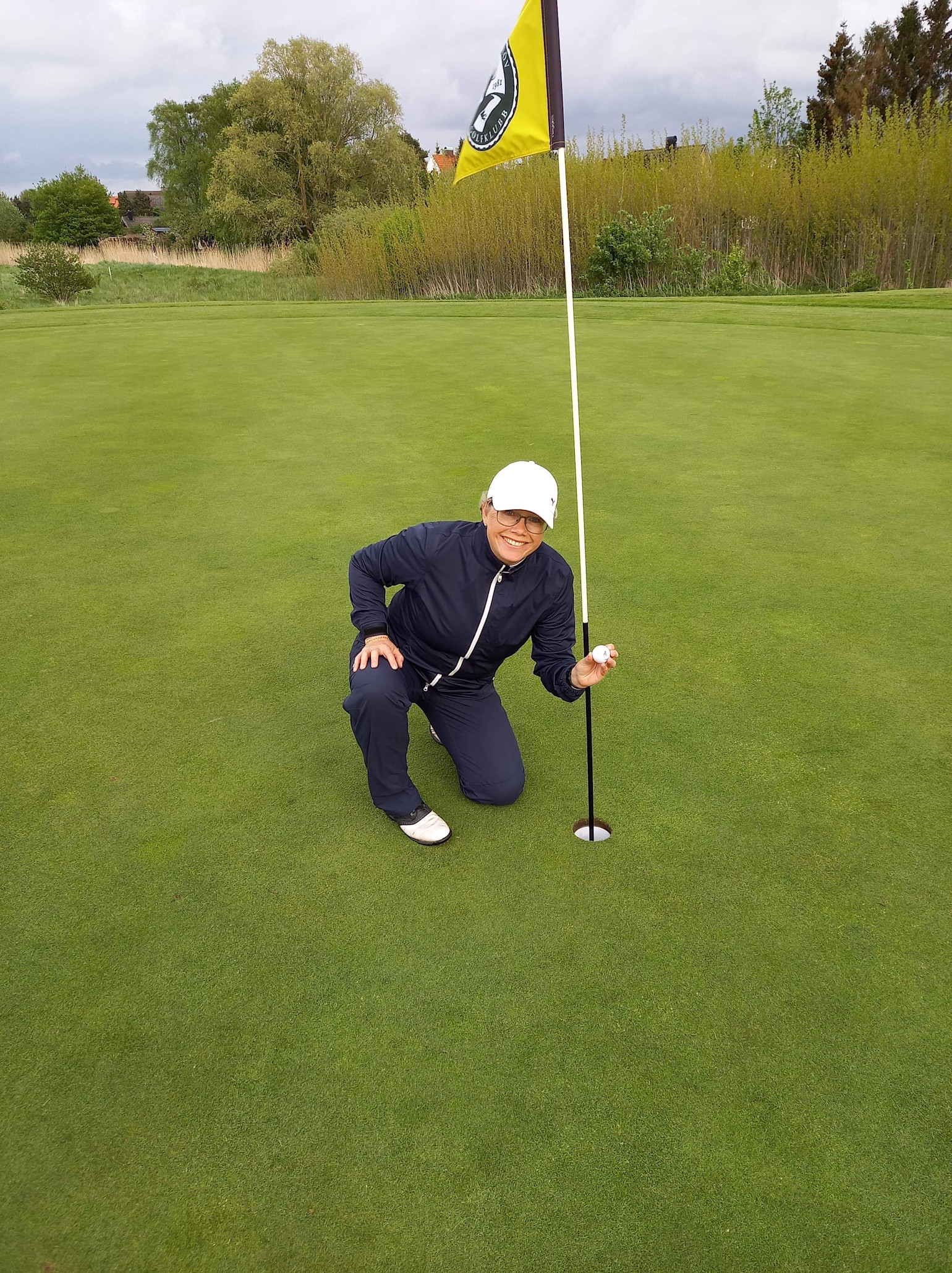 Person knäböjer bredvid golfhål, håller en golfboll och ler, med golfbanans flagga och grönska i bakgrunden.