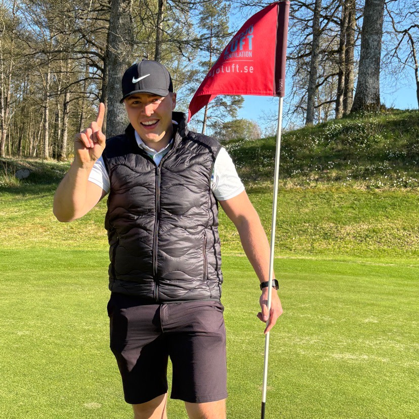 Man i svart keps och väst står på en golfbana, håller i en flaggstång och visar ettan med fingret.