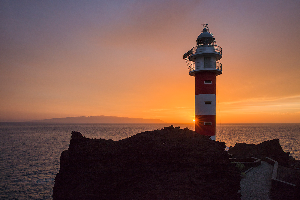 Fyrtorn med rödvita r änder står på klippa vid havet under en färgstark solnedgång.