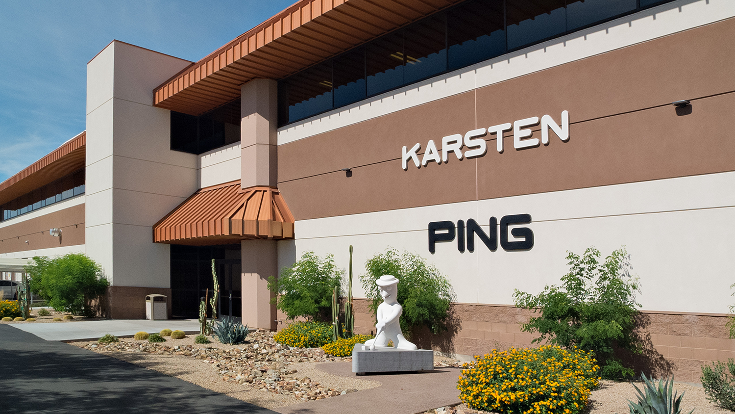 Företagsbyggnad med texten "KARSTEN PING" på fasaden, omgiven av ökenväxter och en vit staty vid ingången.