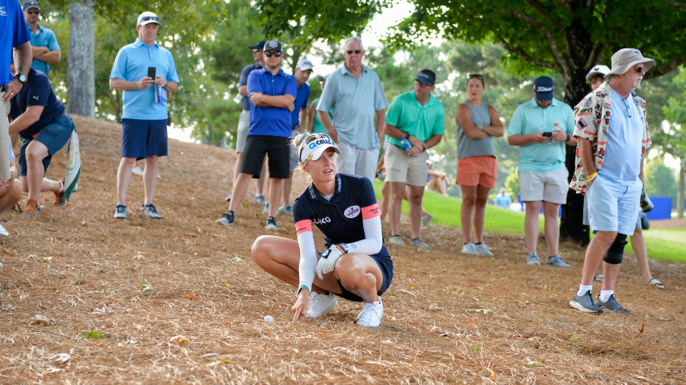 Golfspelare hukar sig och pekar mot en golfboll på marken, omgiven av åskådare i avslappnad miljö utomhus.