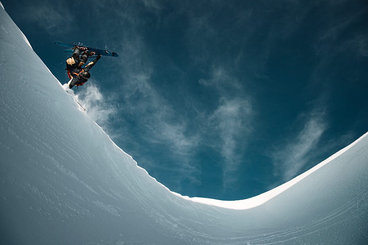En skidåkare gör en bakåtvolt i luften ovanför en snötäckt bergskant under en klarblå himmel.