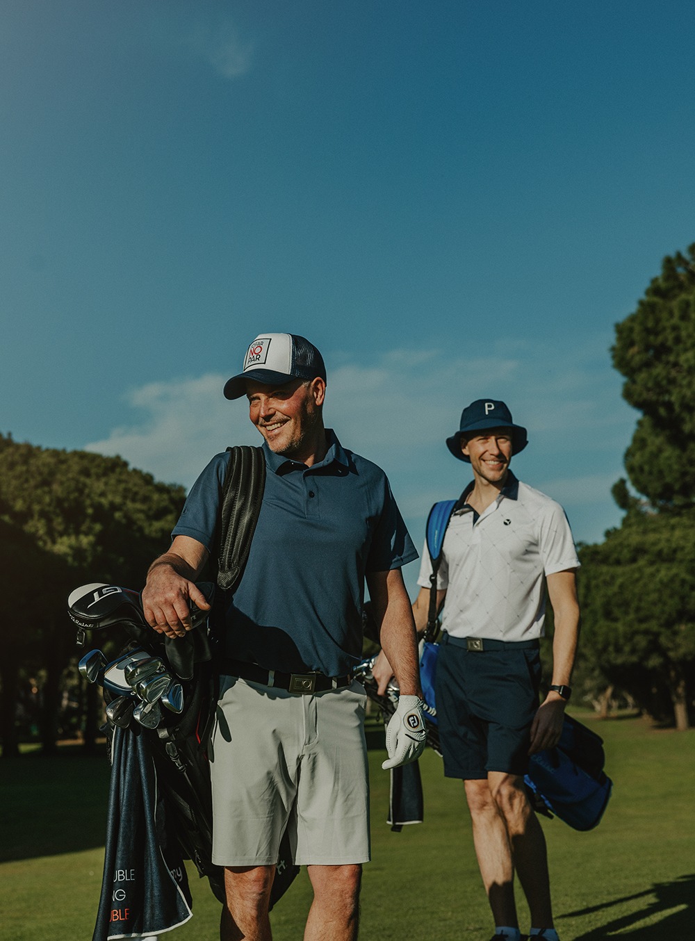 Två män bär golfbagar och ler medan de går på en golfbana under klarblå himmel.