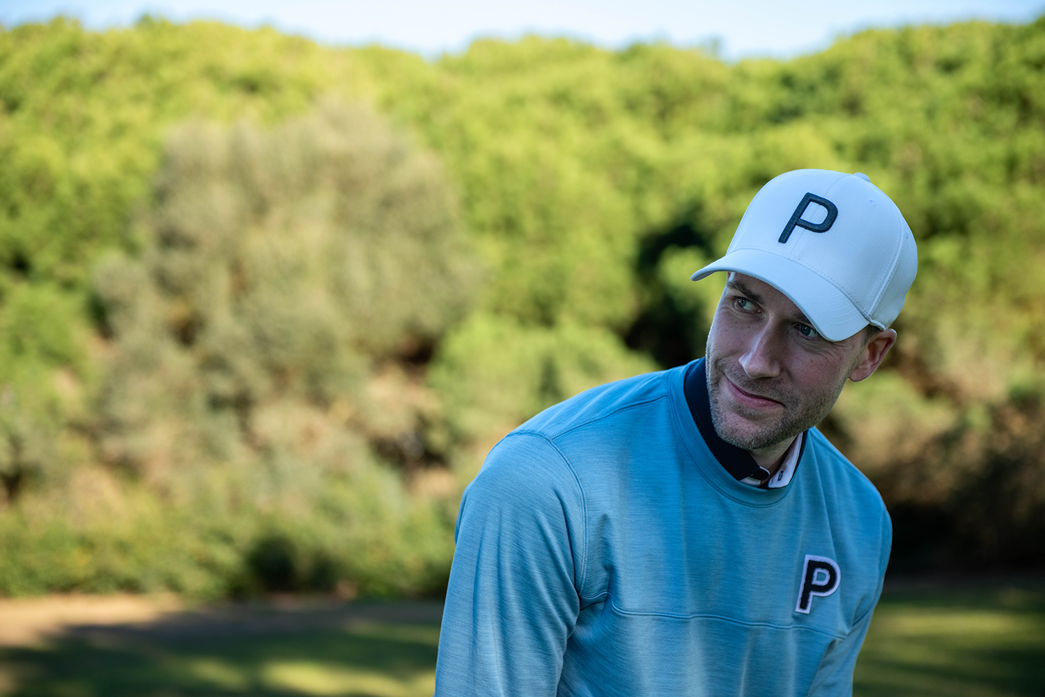 Man med vit keps och blå tröja, båda med bokstaven P, står utomhus framför grönskande träd.