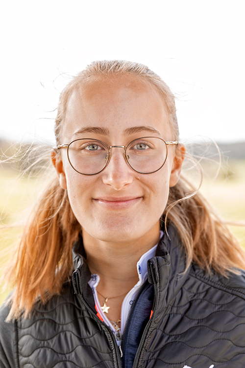 Ung kvinna med glasögon och blont hår i tofsar ler utomhus, iförd mörk jacka och halsband med stjärnhänge.
