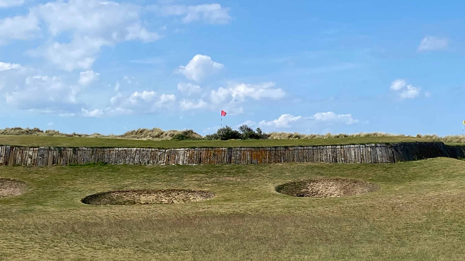 Golfbana med sandbunkrar i förgrunden, trävägg och en röd flagga på greenen under blå himmel.