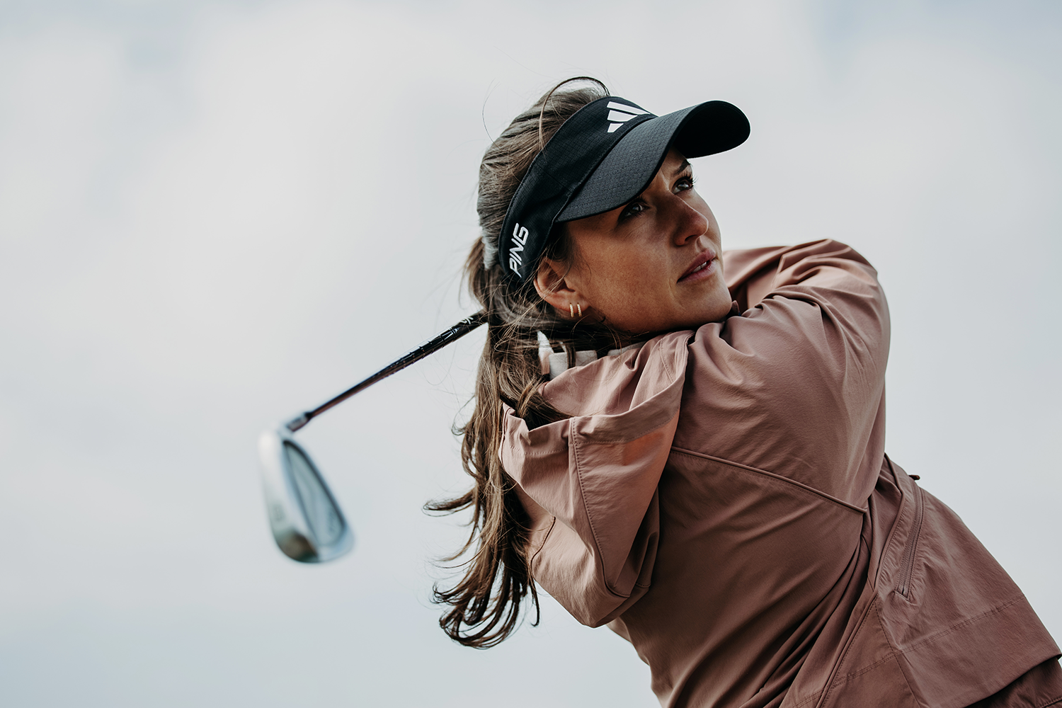 Kvinna i rosa jacka och svart keps svingar en golfklubba utomhus mot ljus bakgrund.