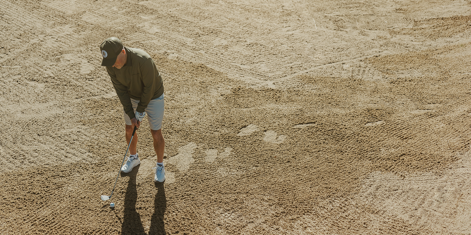 A man in a cap and sportswear is hitting a golf ball with a club from a sand bunker on a golf course.