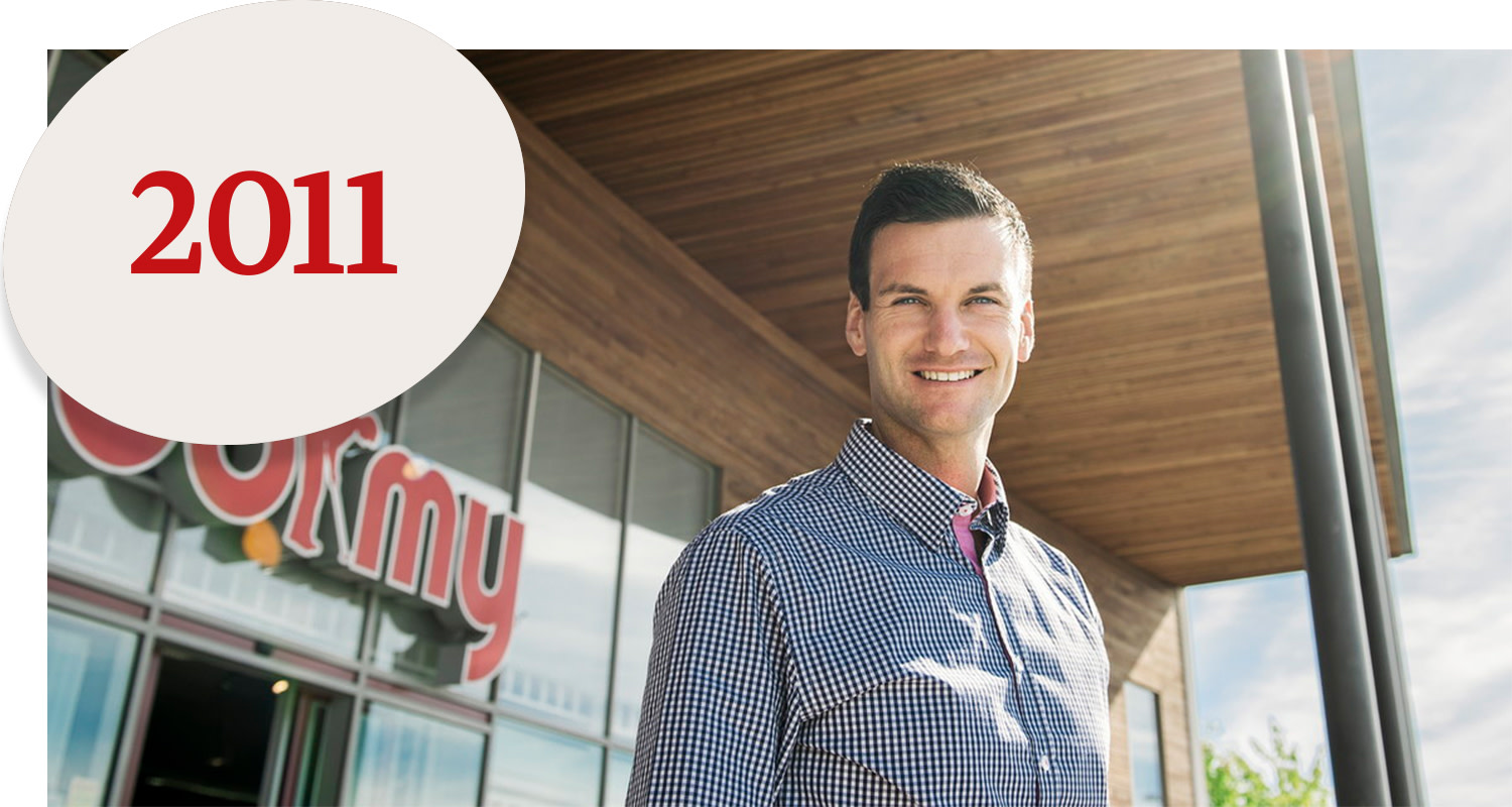 Smiling man in a checked shirt stands outside a building with a wooden facade; a large sign with the text "2011" in red is visible in the corner.