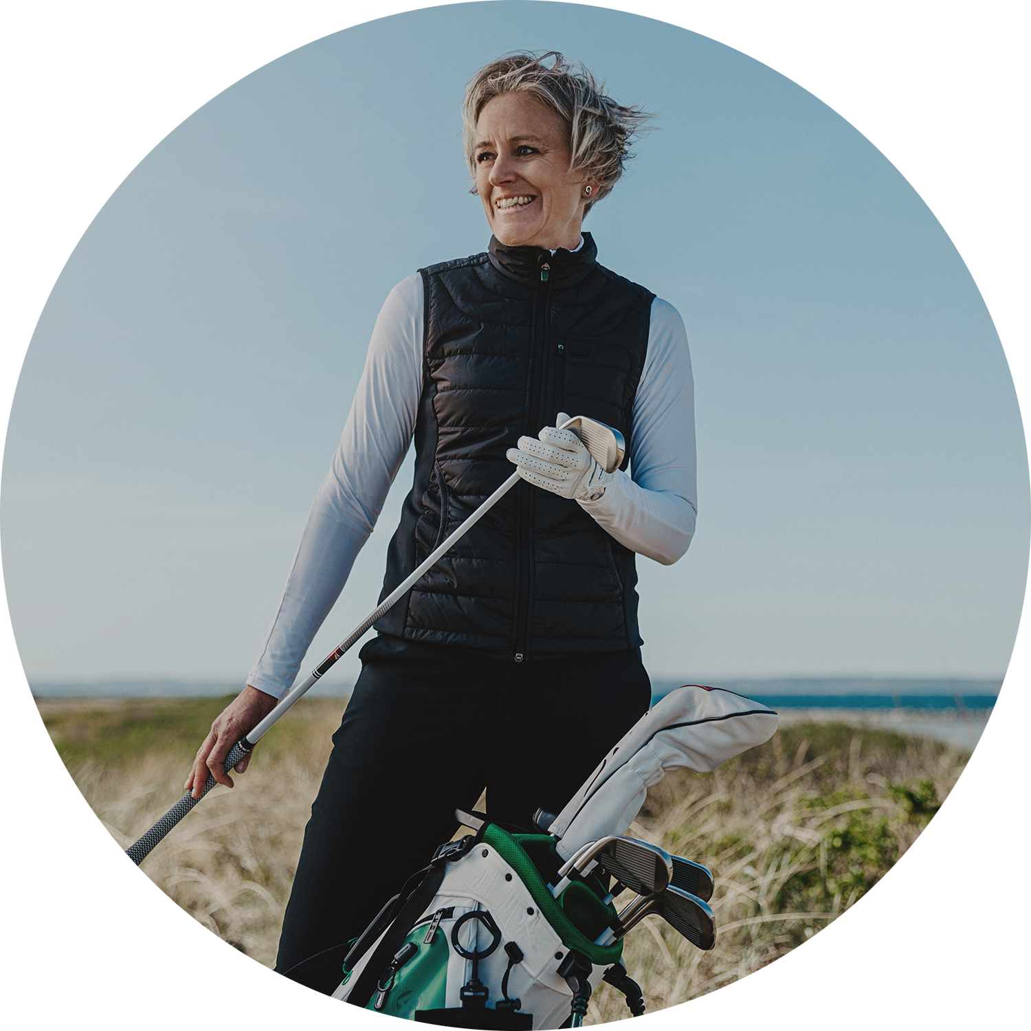 Kvinna med kort hår håller en golfklubba och står bredvid en golfbag utomhus på en golfbana.