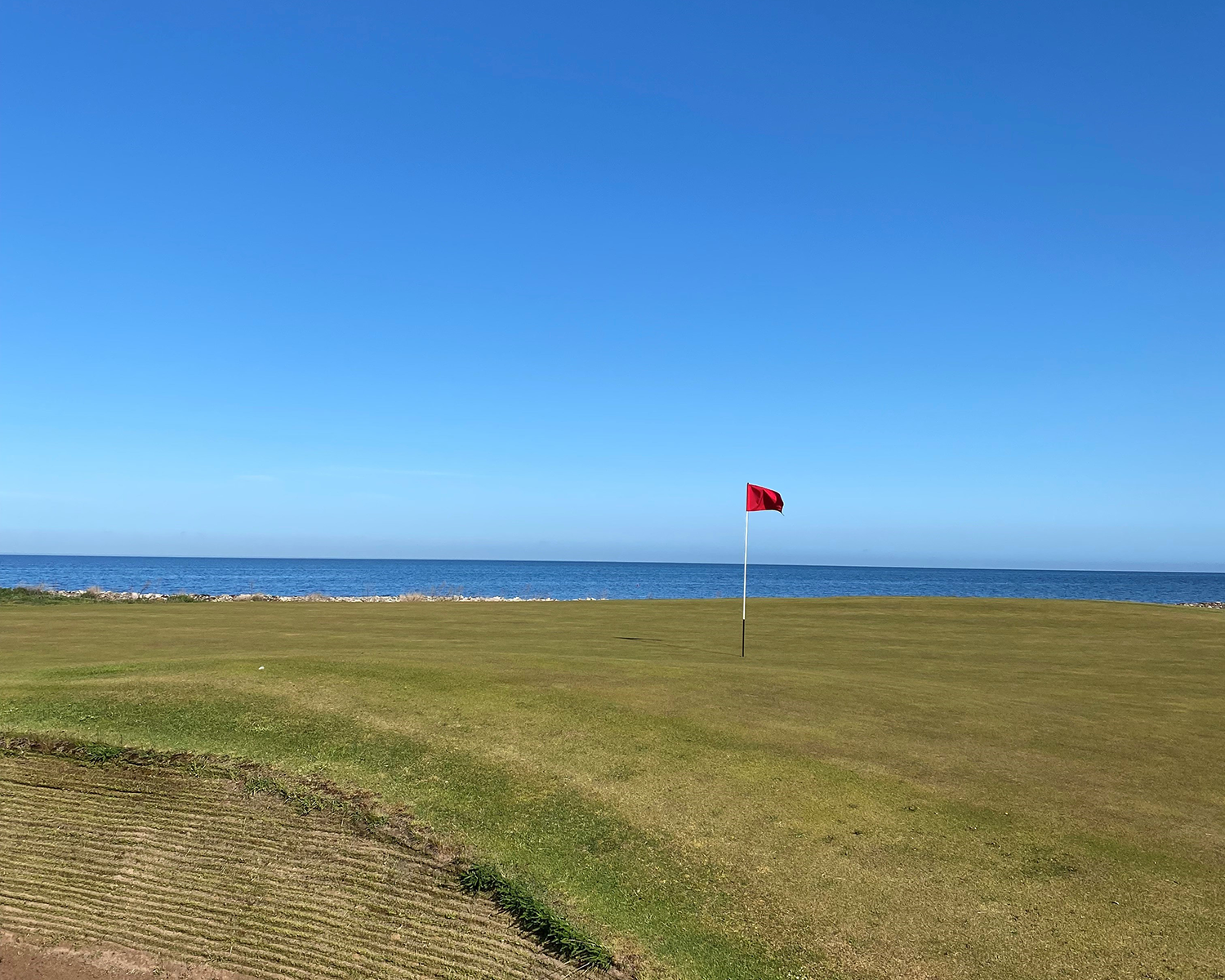 Golfhål med röd flagga på green, havet i bakgrunden och klarblå himmel ovanför.
