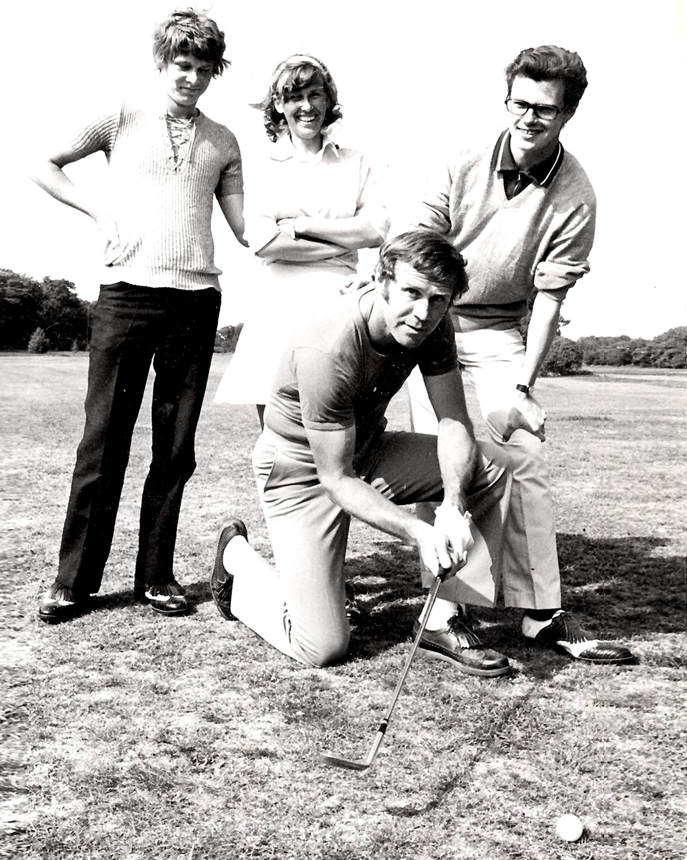 Fyra personer poserar på en golfbana, en man knäböjer och siktar med en golfklubba mot en golfboll, de andra står bakom och ler.