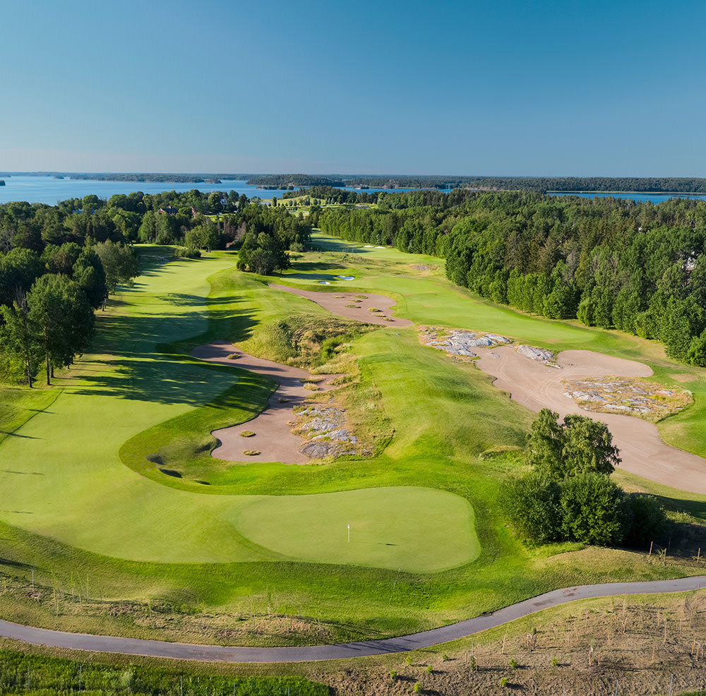 Golfbana med gröna fairways, sandbunkrar och omgivande skog, sjöar syns i bakgrunden under klarblå himmel.