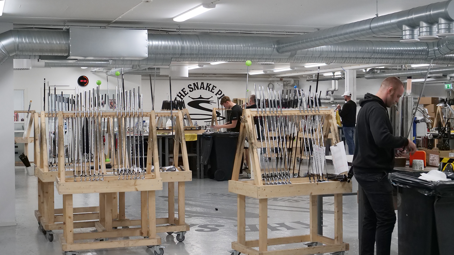 Flera personer arbetar i en fabriksmiljö med golfklubbor placerade på träställningar, ventilationsrör syns i taket.