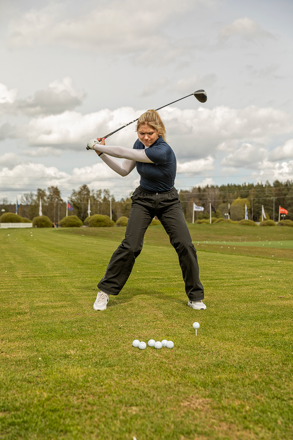Kvinna svingar en golfklubba på driving range, flera golfbollar ligger på gräset framför henne under molnig himmel.