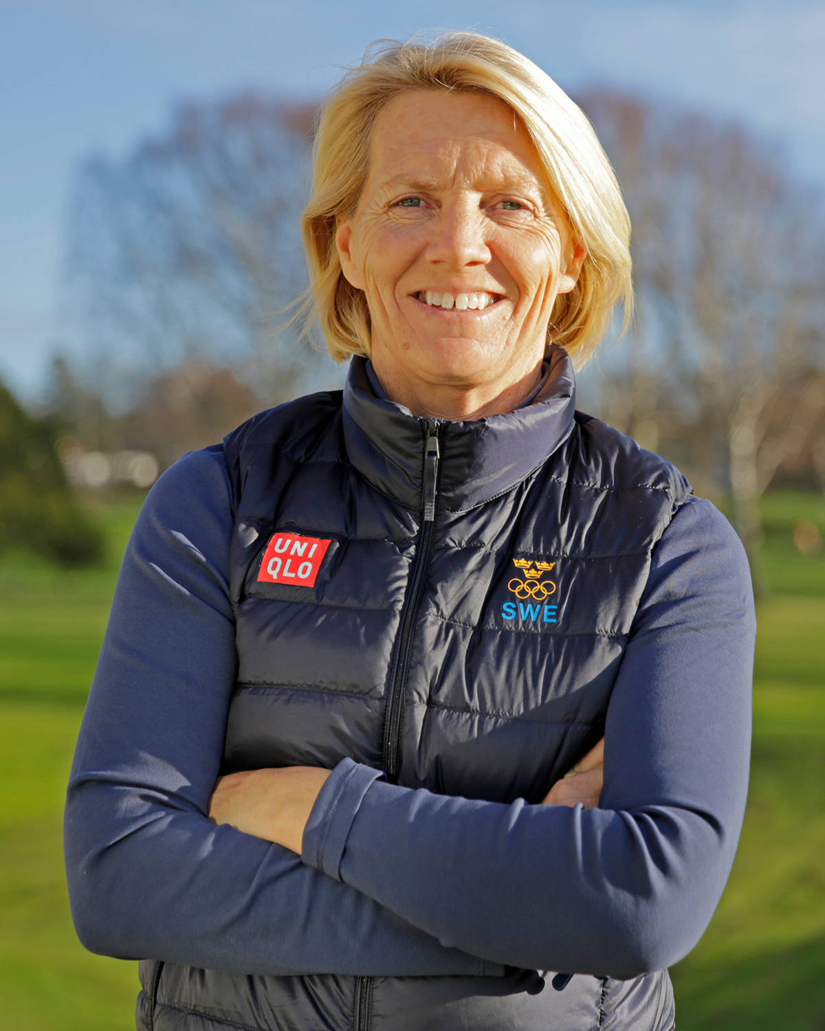 Kvinna med blont hår står utomhus med armarna i kors, bär blå väst med UNIQLO och svenska OS-loggan.