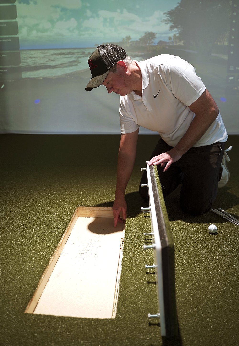 Man i vit pikétröja och keps undersöker en öppnad sektion av konstgräsmatta inomhus, med golfboll och klubbor bredvid.