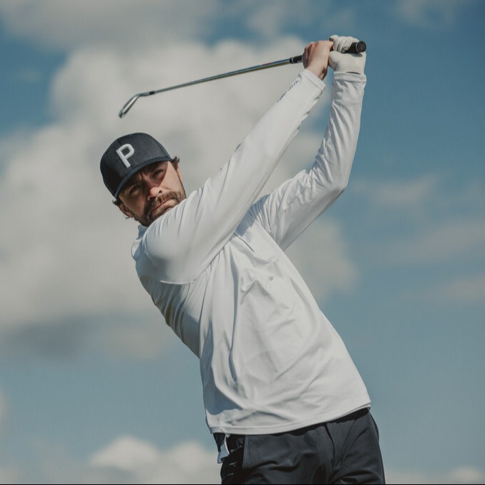 Man med skägg svingar en golfklubba utomhus, bär vit tröja och mörk keps med bokstaven P mot blå himmel.