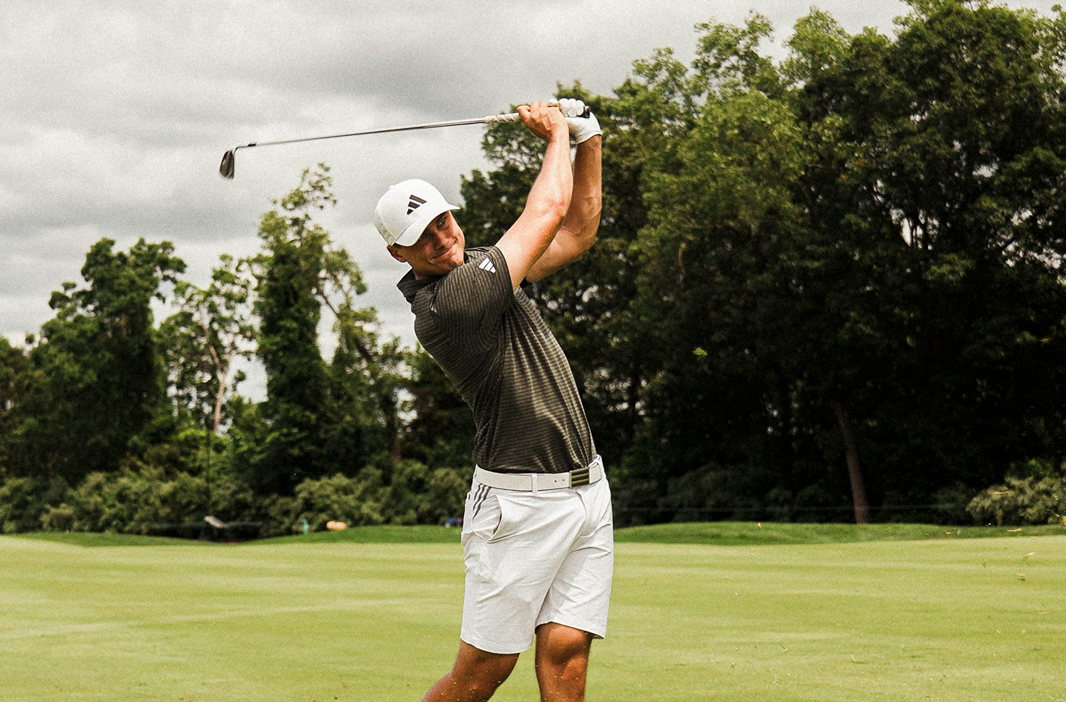 Golfspelare svingar en klubba på en grön golfbana, omgiven av träd under en molnig himmel.