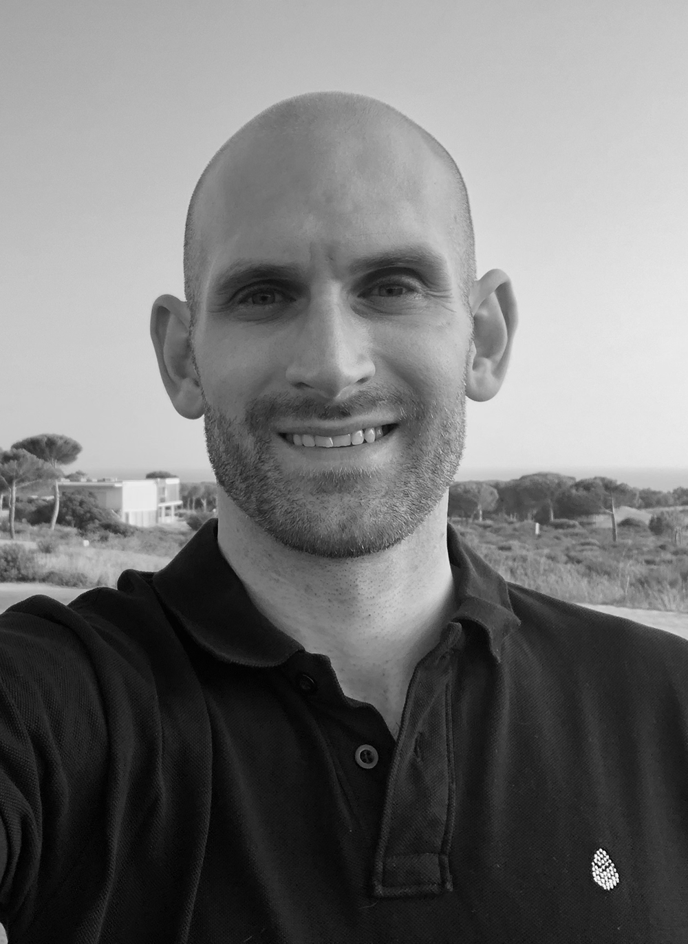 Skäggig man med rakat huvud ler och tar en selfie utomhus, med landskap och byggnader i bakgrunden.