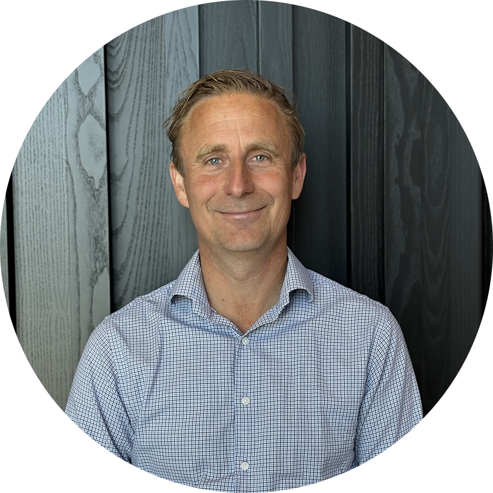 Man ler mot kameran, sitter framför en vägg med mörka och ljusa träpaneler, bär en ljusblå rutig skjorta.