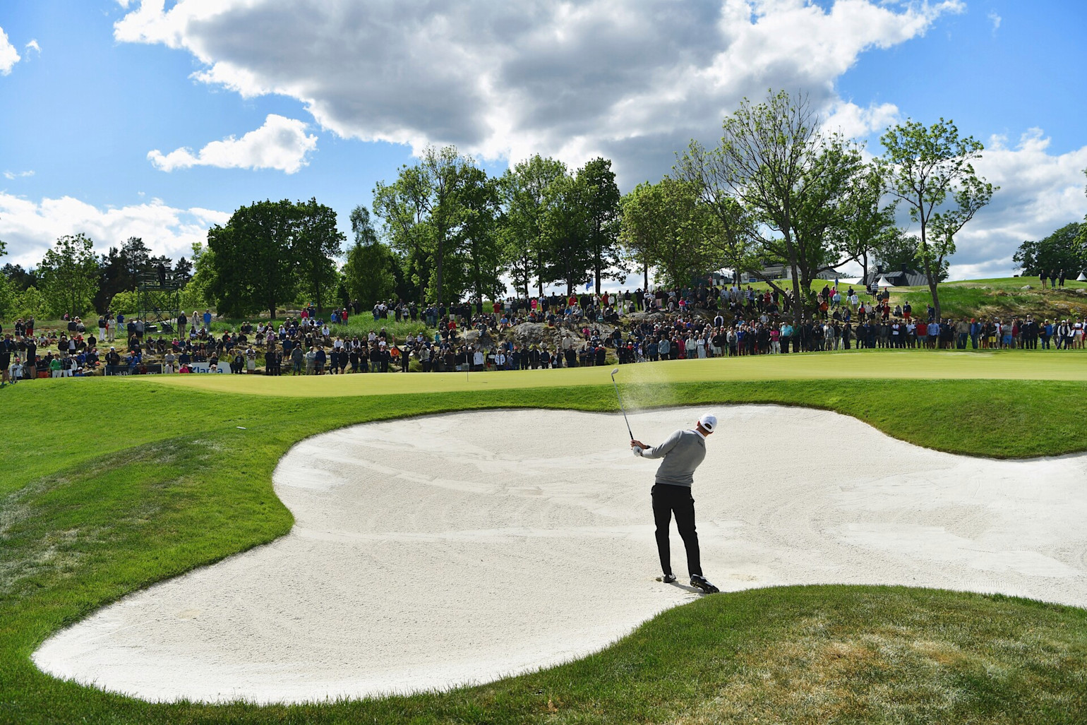 Golfspelare slår ett slag ur en bunker mot greenen medan en stor publik tittar på i bakgrunden under en solig dag.