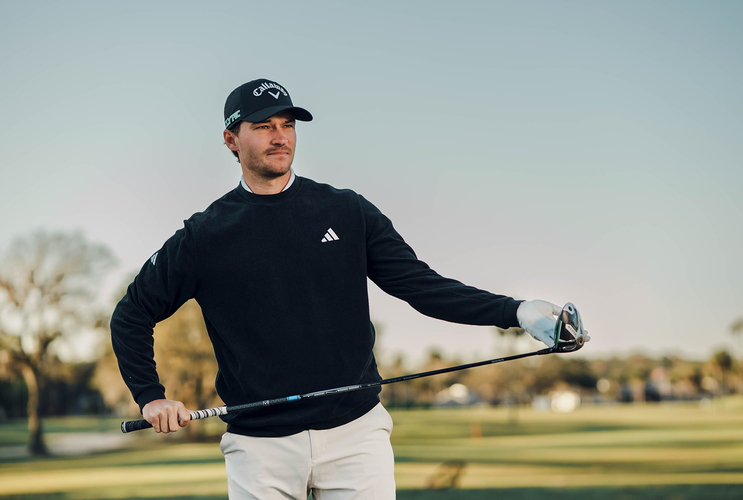 Man i svart tröja och keps håller en golfklubba på en golfbana under klar himmel.
