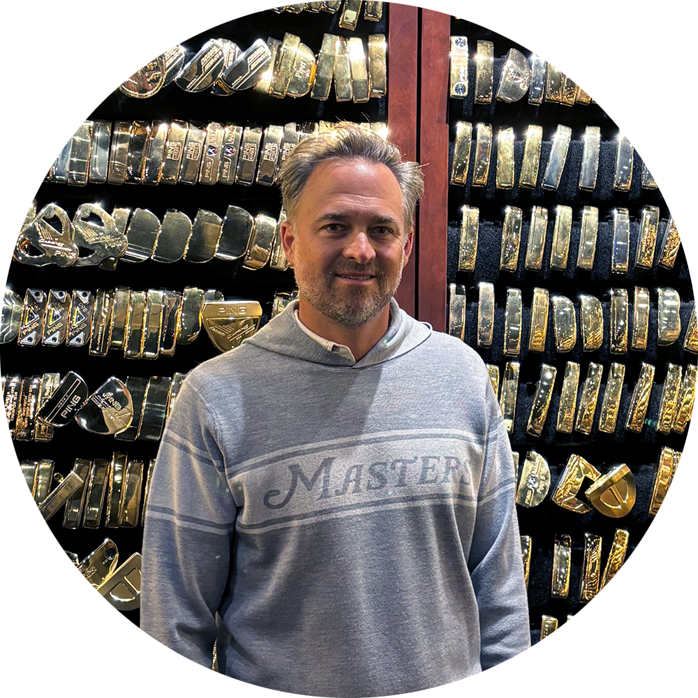 Man i grå huvtröja med texten "Masters" står framför en vägg fylld med glänsande golfklubbor och putters.