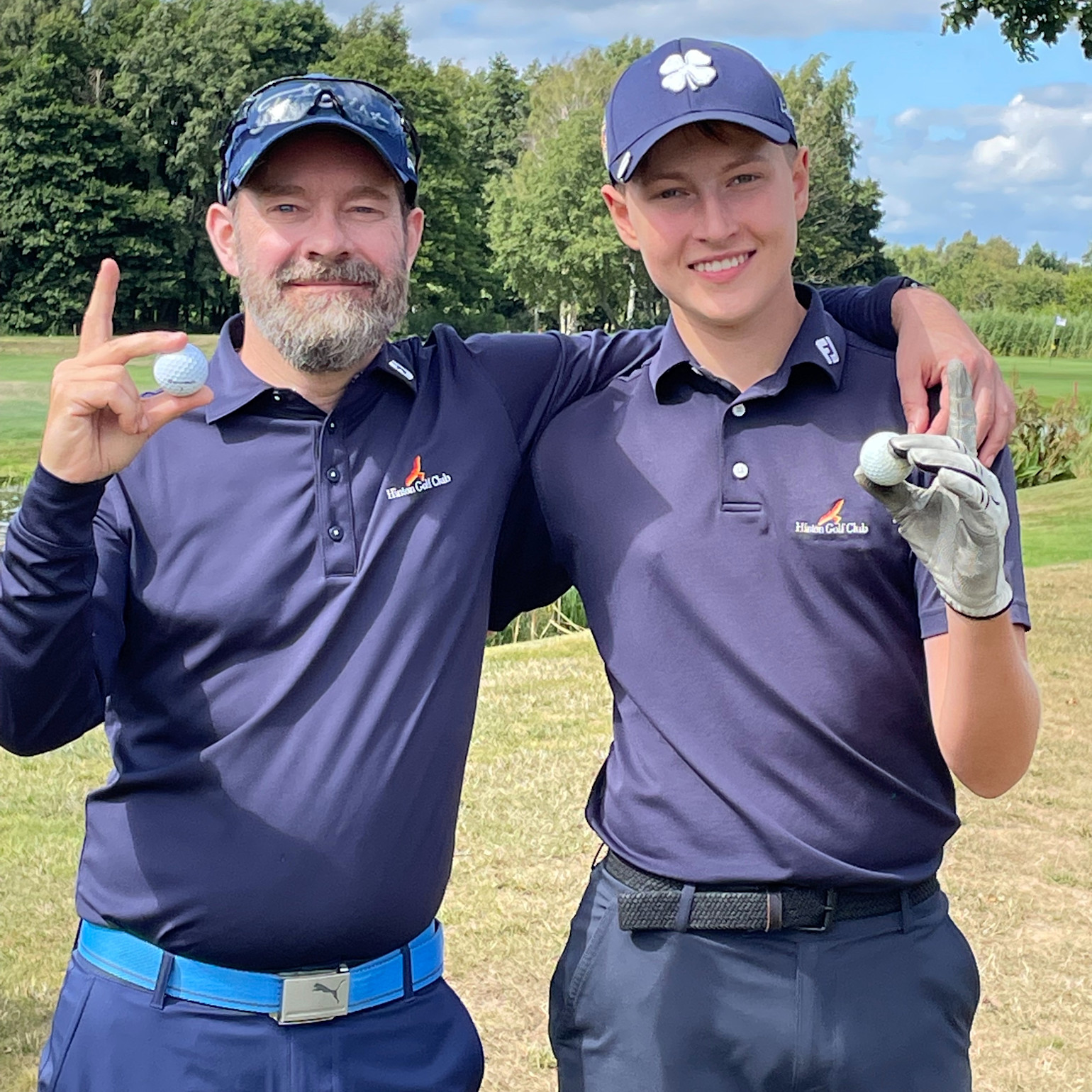 Två män i mörkblå Hilson Golf Club-tröjor håller golfbollar och ler på en golfbana med grönska i bakgrunden.