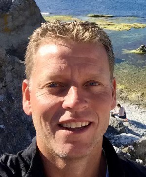 Man ler mot kameran utomhus vid en klippig strand med havet och grönalger i bakgrunden.