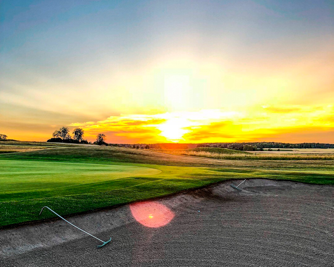 Golfbana med sandbunker och krattor i förgrunden, solnedgång över gröna fält och träd i bakgrunden.