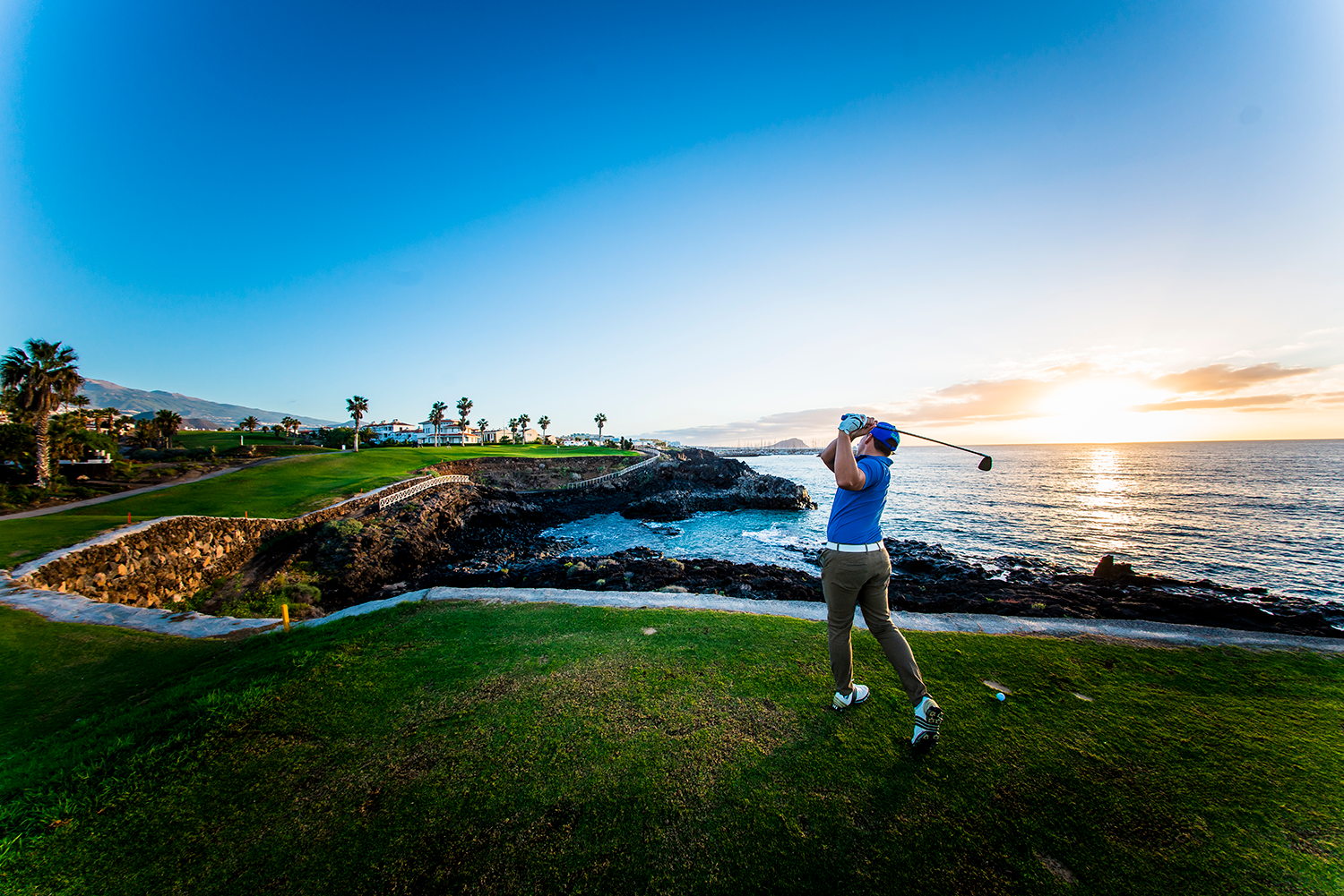 Golfspelare svingar en klubba vid havet på en grön golfbana i solnedgången, med palmer och berg i bakgrunden.