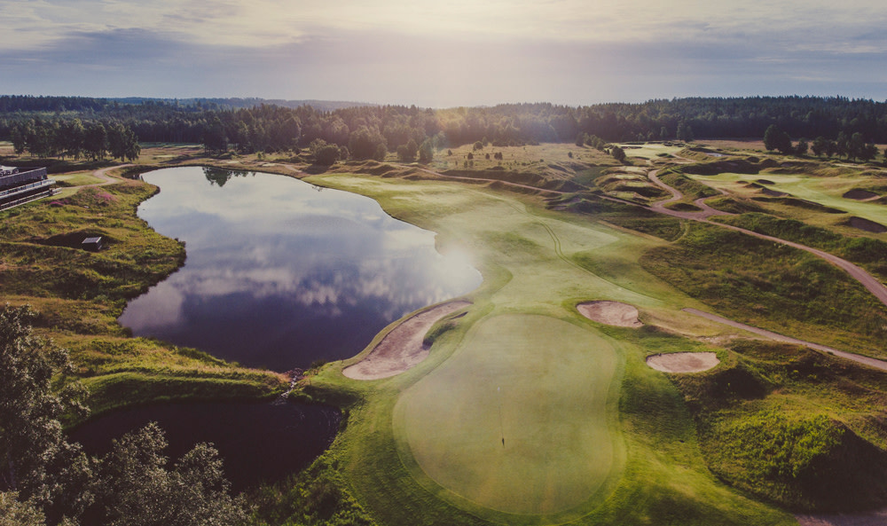 Golfbana med greener och sandbunkrar bredvid en stor sjö, omgiven av grönska och skog i solnedgång.