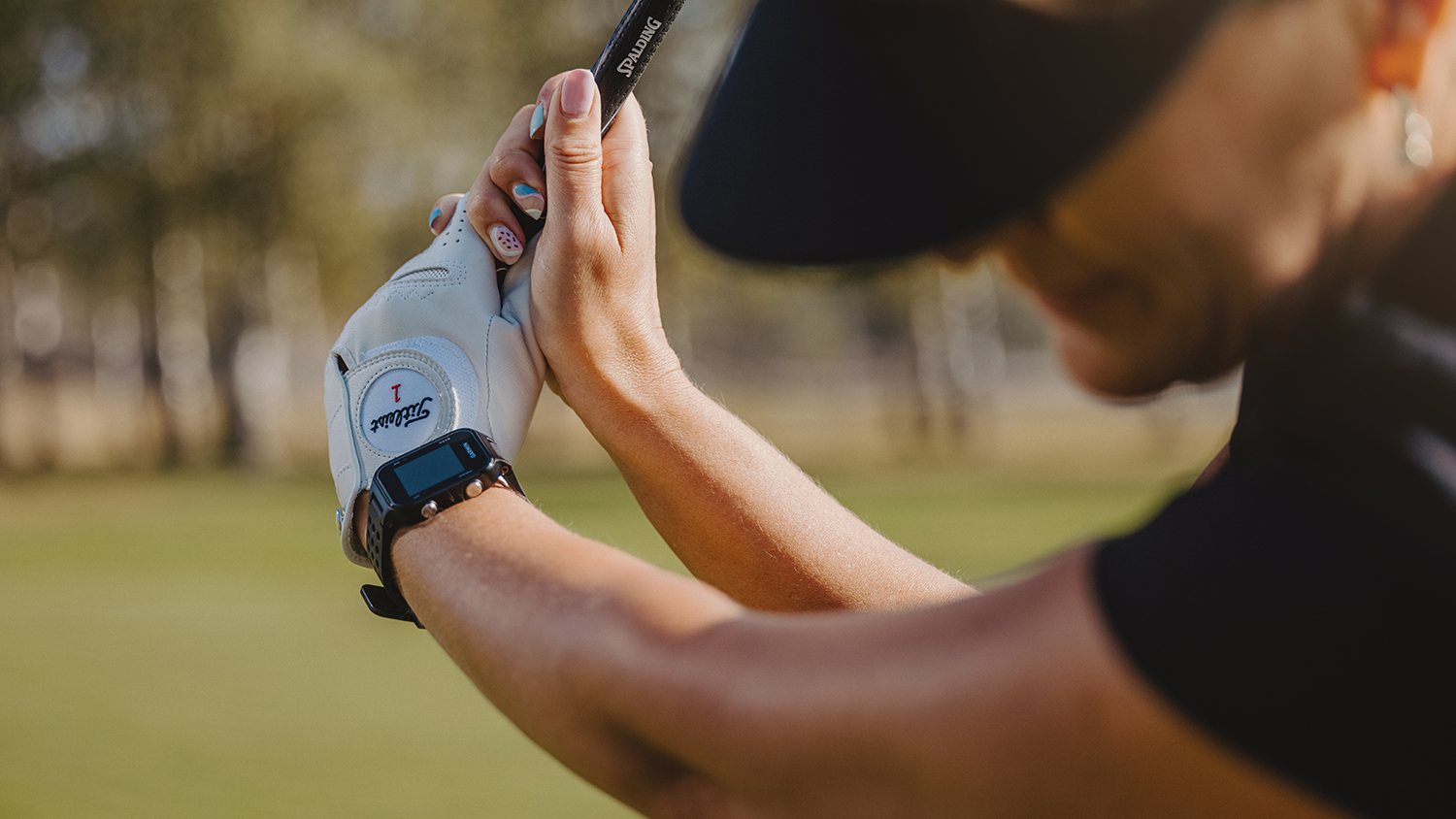 Närbild på en person som svingar en golfklubba, bär golfhandske från Titleist och sportklocka, utomhus på golfbana.