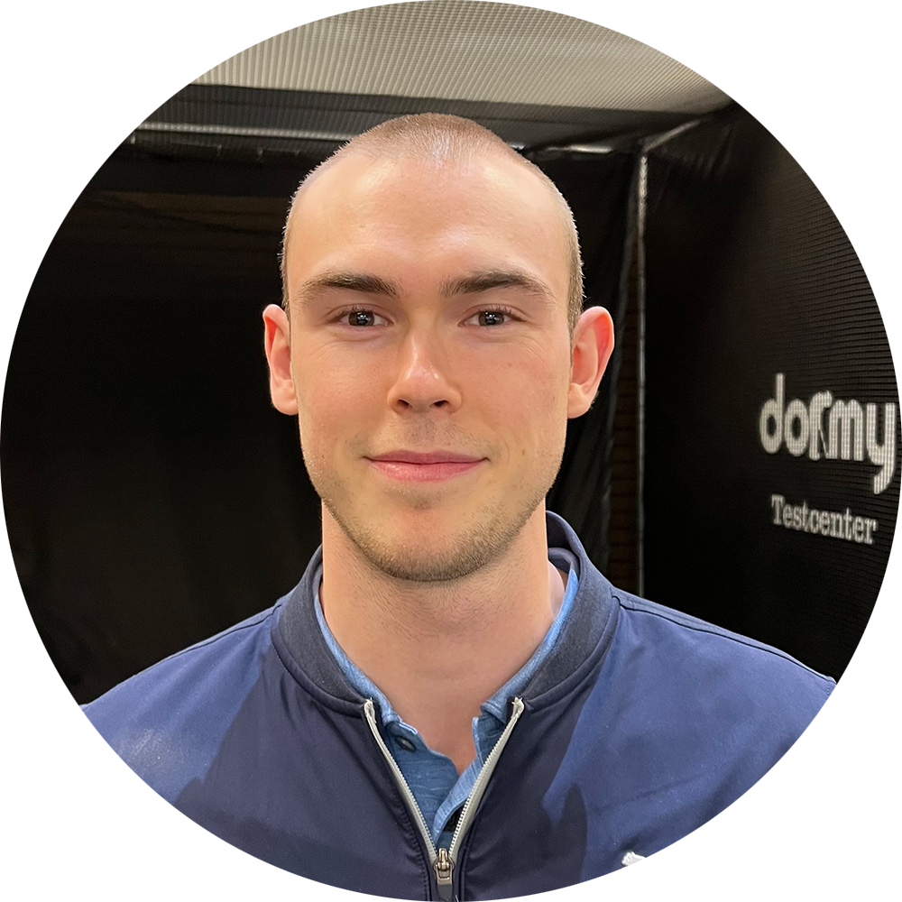 Ung man med kortklippt hår ler mot kameran, står framför en svart vägg med texten "Dormy Testcenter".