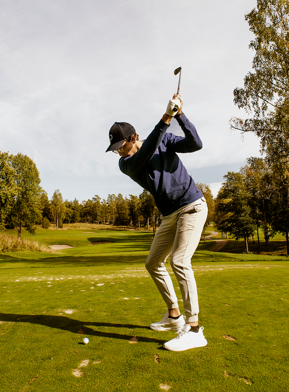 Man i keps och glasögon svingar en golfklubba på en grön golfbana omgiven av träd under klar himmel.