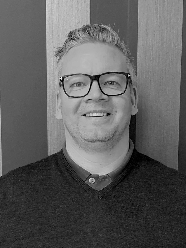 Man med glasögon ler mot kameran, står framför en vägg med breda, vertikala ränder i olika nyanser.