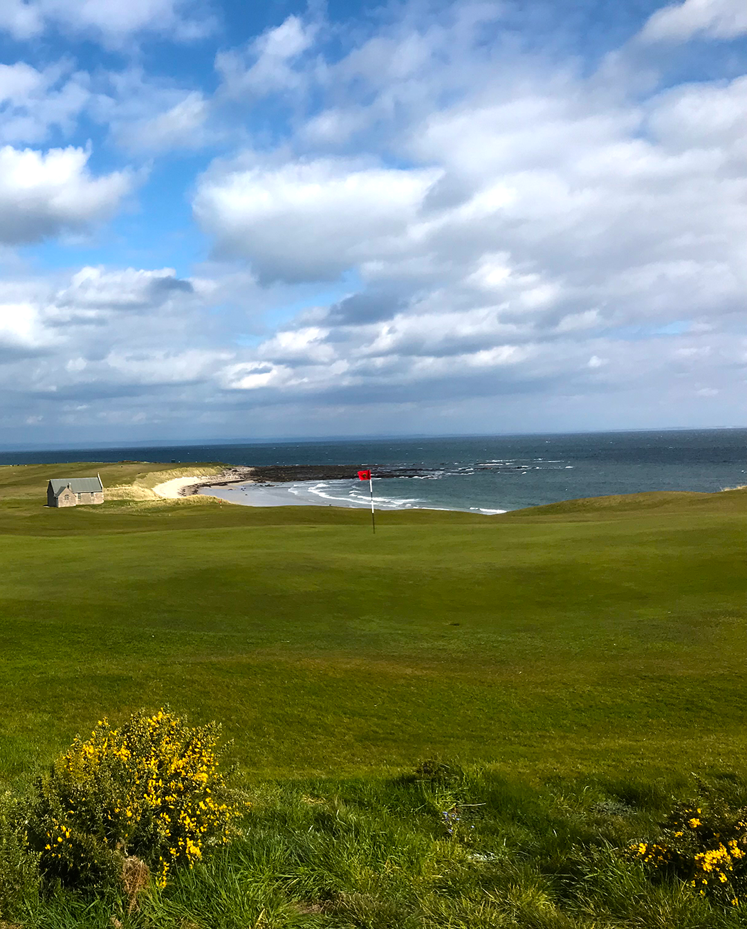 Golfbana med en röd flagga på green, havet i bakgrunden och ett litet stenhus vid stranden under en molnig himmel.